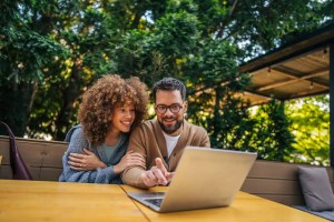 Casal sorridente em área externa, conversando e trabalhando com notebook em mesa de madeira sob um guarda-sol, cercados por árvores e vegetação ao fundo.