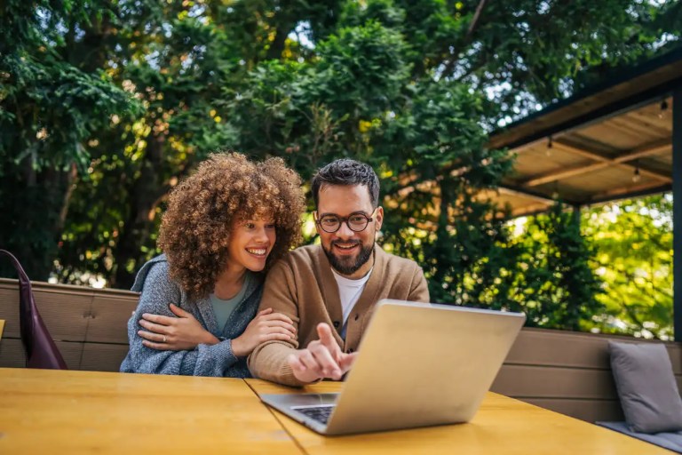 Casal sorridente em área externa, conversando e trabalhando com notebook em mesa de madeira sob um guarda-sol, cercados por árvores e vegetação ao fundo.