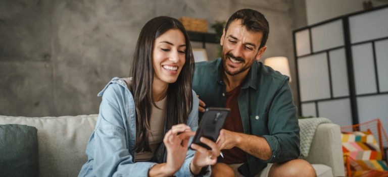Casal sorridente sentado no sofá usando smartphone juntos, desfrutando de momentos felizes em casa.
