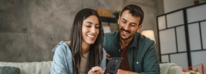 Casal sorridente sentado no sofá usando smartphone juntos, desfrutando de momentos felizes em casa.