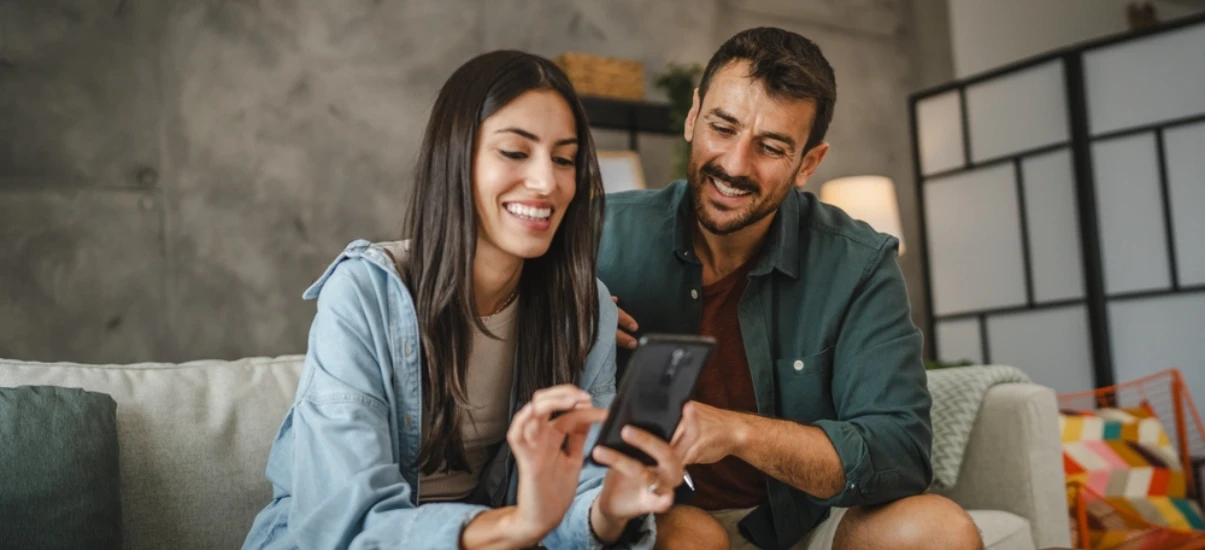 Casal sorridente sentado no sofá usando smartphone juntos, desfrutando de momentos felizes em casa.