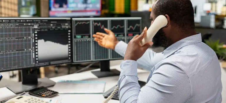 Homem de terno trabalhando em múltiplos monitores com gráficos financeiros, analisando dados de mercado e realizando operações na bolsa de valores.