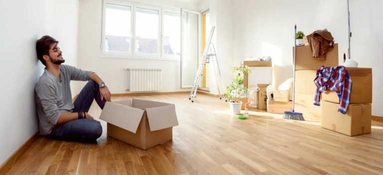 Homem jovem feliz sentado no piso de um apartamento vazio, em processo de mudança, com caixas de papelão, escada, plantas e móveis empacotados, simbolizando organização e mudança residencial.