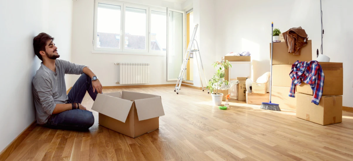 Homem jovem feliz sentado no piso de um apartamento vazio, em processo de mudança, com caixas de papelão, escada, plantas e móveis empacotados, simbolizando organização e mudança residencial.