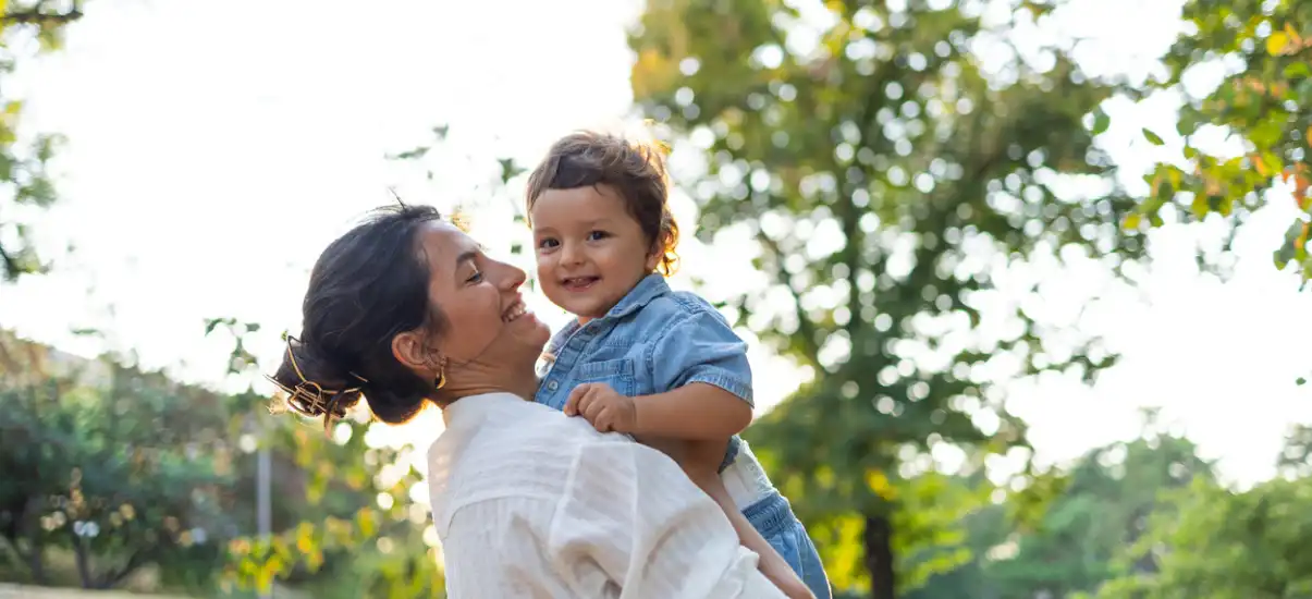 Mulher segurando uma criança sorridente ao ar livre, cercada por árvores verdes e céu claro, em um ambiente natural e alegre.
