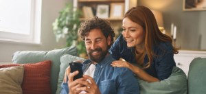 Casal sorridente no sofá da sala, com homem segurando o celular e a mulher ao lado, sorrindo e inclinada para conversar, em ambiente aconchegante com planta ao fundo.