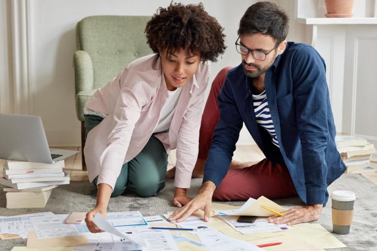 Homem e mulher sentados, com diversos papéis, enquanto fazem contas para seu equilíbrio financeiro.