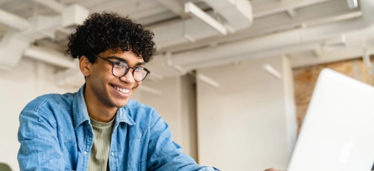 Jovem rapaz com cabelo cacheado, usando óculos e camiseta verde, sentado em ambiente de escritório, sorrindo ao trabalhar no computador.