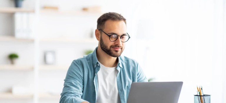 Homem de óculos usando laptop em um ambiente de escritório bem iluminado, com expressão concentrada, ideal para trabalhos remotos e produtividade.