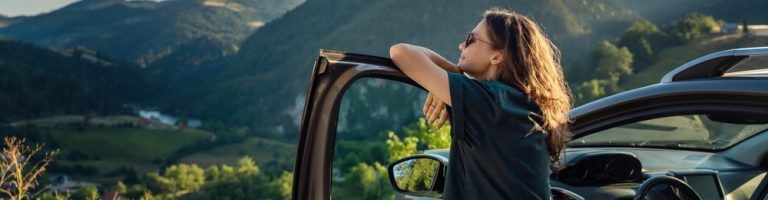 Mulher com óculos escuro encostado em um carro olhando a paisagem nas montanhas