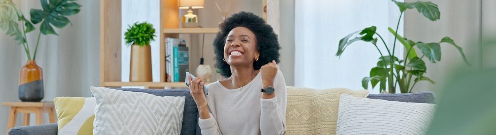Mulher feliz sentada em um sofá em uma sala iluminada, expressando alegria e satisfação, com sofás confortáveis, plantas e objetos decorativos ao fundo.