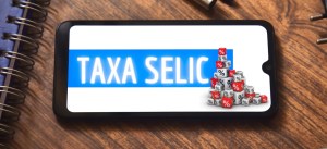 Imagem de um smartphone exibindo a expressão 'Taxa Selic' com um gráfico de blocos representando porcentagens, ao lado de uma mesa de madeira com notebooks e canetas.