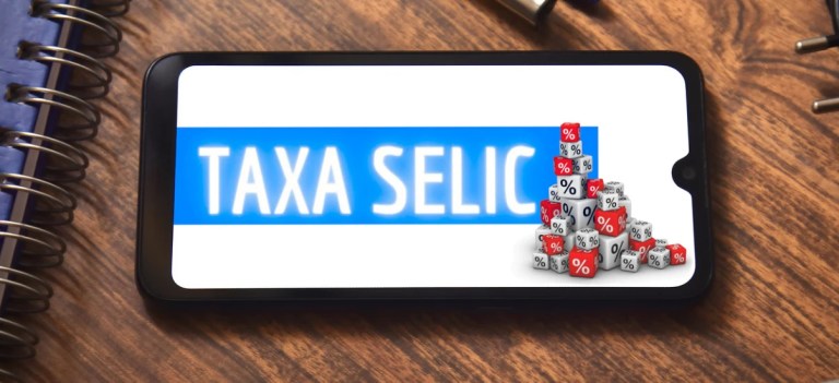 Imagem de um smartphone exibindo a expressão 'Taxa Selic' com um gráfico de blocos representando porcentagens, ao lado de uma mesa de madeira com notebooks e canetas.