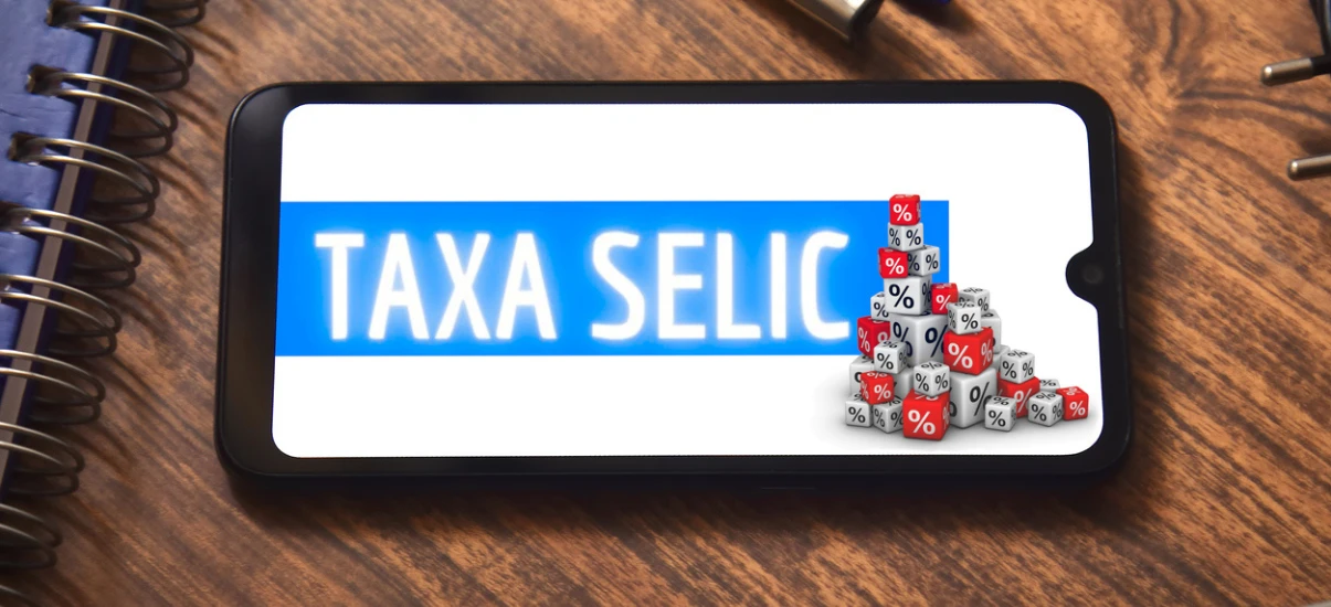 Imagem de um smartphone exibindo a expressão 'Taxa Selic' com um gráfico de blocos representando porcentagens, ao lado de uma mesa de madeira com notebooks e canetas.