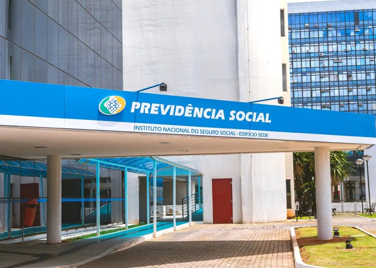 Fachada do prédio com a placa “Previdência Social”, do Instituto Nacional do Seguro Social, com área externa e cobertura de vidro ao lado.