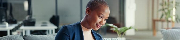 Mulher com cabelo curto e vestido azul, sorrindo enquanto utiliza um tablet em um ambiente de escritório moderno. Dicas para produtividade e tecnologia.