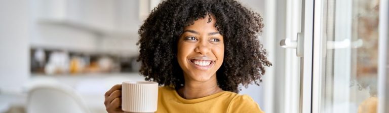 Mulher com cabelo cacheado, sorrindo e segurando uma caneca em um ambiente aconchegante.