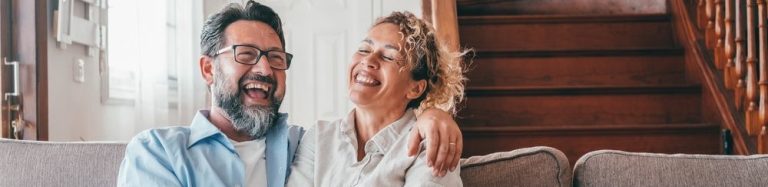 Casal sorridente se divertindo no sofá de casa, demonstrando amor e alegria em um ambiente aconchegante.