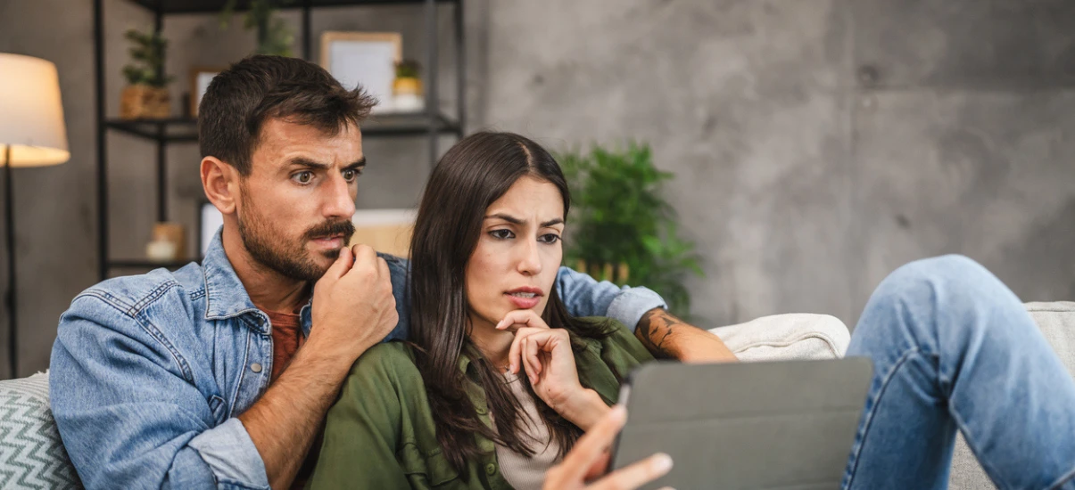 Casal assistindo a uma tela de dispositivo eletrônico na sala de estar, demonstrando preocupação e atenção.