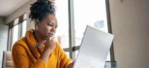 Mulher pensando enquanto analisa informações no laptop, em ambiente de escritório com iluminação natural e janelas grandes.