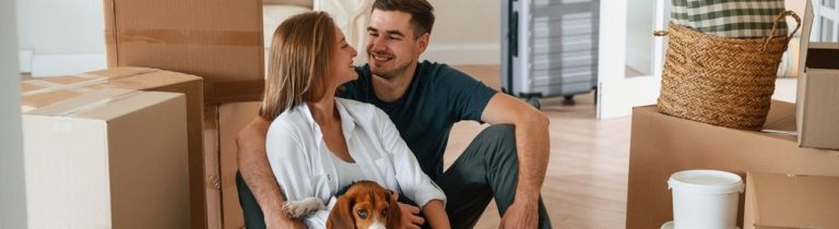 Casal sorridente sentado no chão de uma sala com caixas de mudança e um cachorro. Eles parecem felizes e acolhedores em um novo lar.