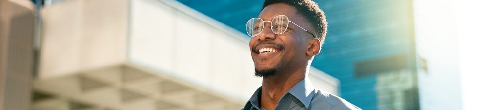 Homem sorridente caminhando em área urbana moderna, usando camisa azul e óculos, transmitindo confiança e felicidade.