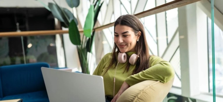 Mulher jovem usando fones de ouvido e trabalhando no notebook em ambiente corporativo, sentada com postura relaxada e expressão sorridente