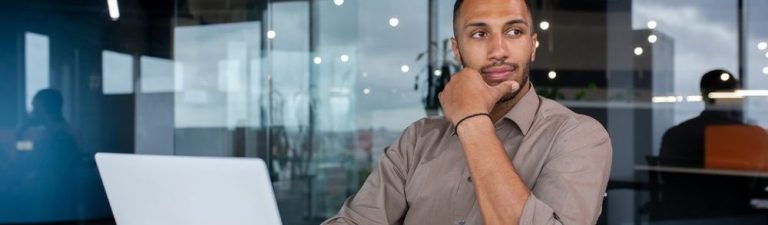 Homem pensativo sentado em um escritório moderno, trabalhando em um laptop com caderno e celular ao lado.