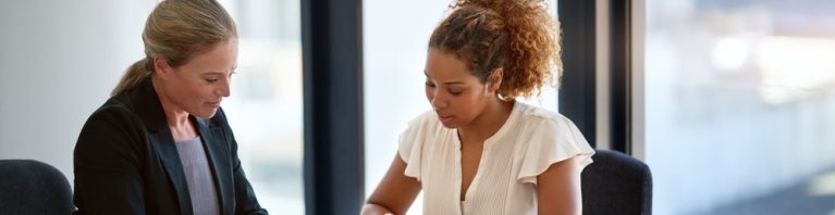 Duas mulheres em um ambiente de trabalho colaborativo, analisando documentos e fazendo anotações em uma mesa cheia de papéis e materiais de escritório.