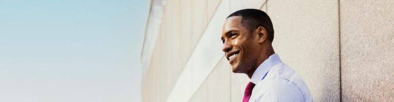 Homem sorridente vestido com camisa e gravata, usando um smartphone em ambiente urbano.