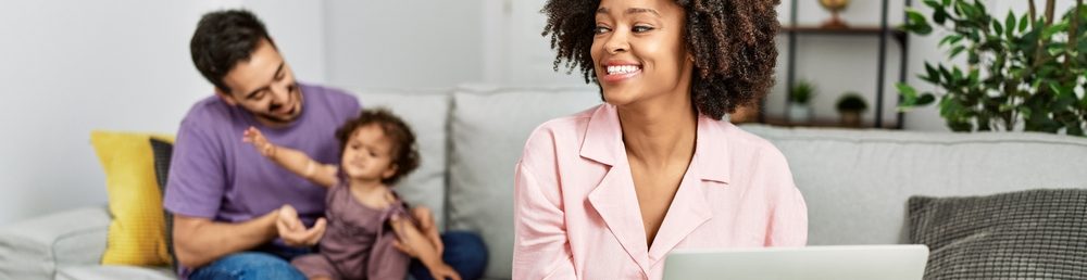 Uma mulher sorridente usando um laptop enquanto um homem brinca com uma criança no sofá.