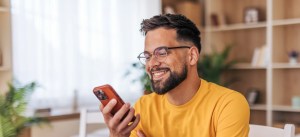 Homem sorridente usando óculos e camiseta amarela, olhando para o celular em ambiente bem iluminado com móveis de madeira e plantas ao fundo.