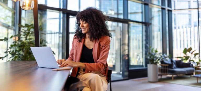 Mulher sorridente trabalhando em home office ou escritório moderno, usando notebook sobre a mesa e olhando para a tela, com janelas amplas e plantas ao fundo.