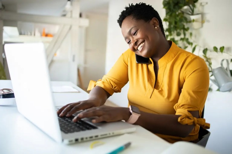 Mulher em frente ao computador trabalhando, ao mesmo tempo em que está falando ao celular