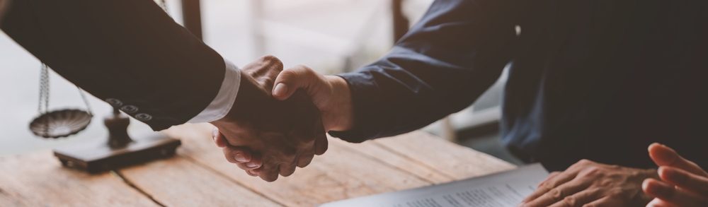 Profissionais apertando as mãos em um ambiente de trabalho, simbolizando um acordo legal com documentos e um martelo de juiz sobre a mesa.