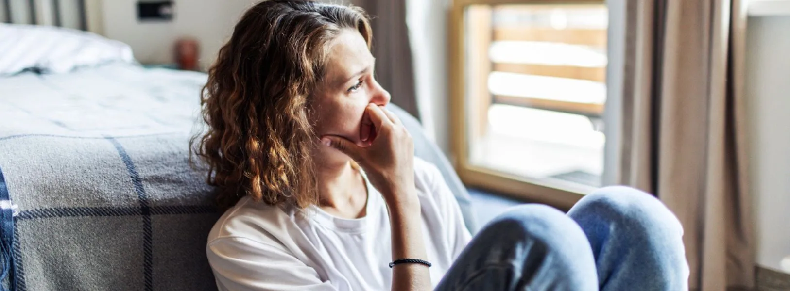 mulher sentada sozinha ao lado da cama no quarto com o rosto triste e olhar pensativo