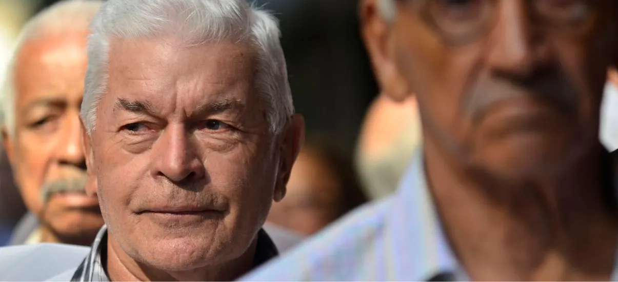 Um grupo de homens mais velhos, focados em um homem central com cabelos brancos, estão de pé ao ar livre. A imagem tem uma profundidade de campo rasa, mantendo o fundo e outros homens desfocados.