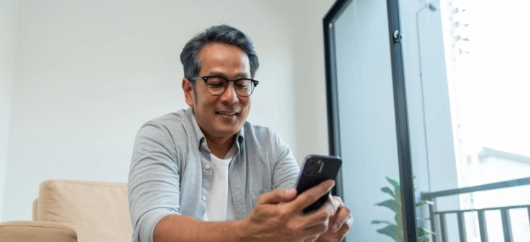 Homem sorridente utilizando celular em ambiente interno bem iluminado, com janela ao fundo e decoração moderna.