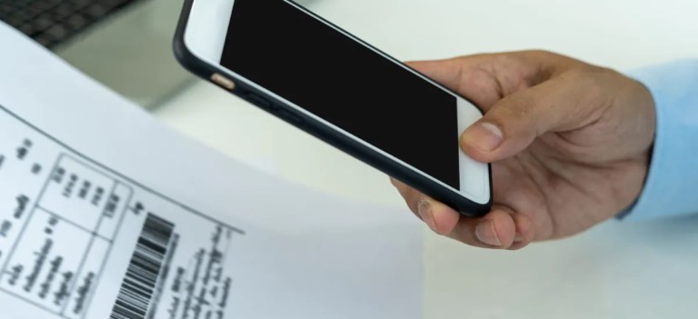 Pessoa segurando um celular próximo a um documento impresso, sugerindo o uso de tecnologia para pagamentos ou verificação de informações.