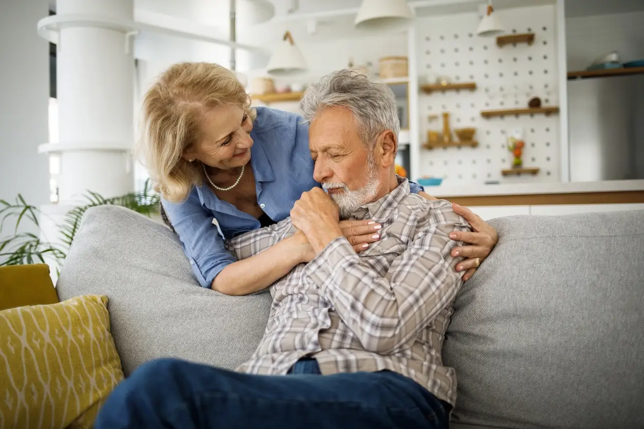 Mulher de meia idade abraça e conforta um seu marido sentado no sofá, em um ambiente acolhedor