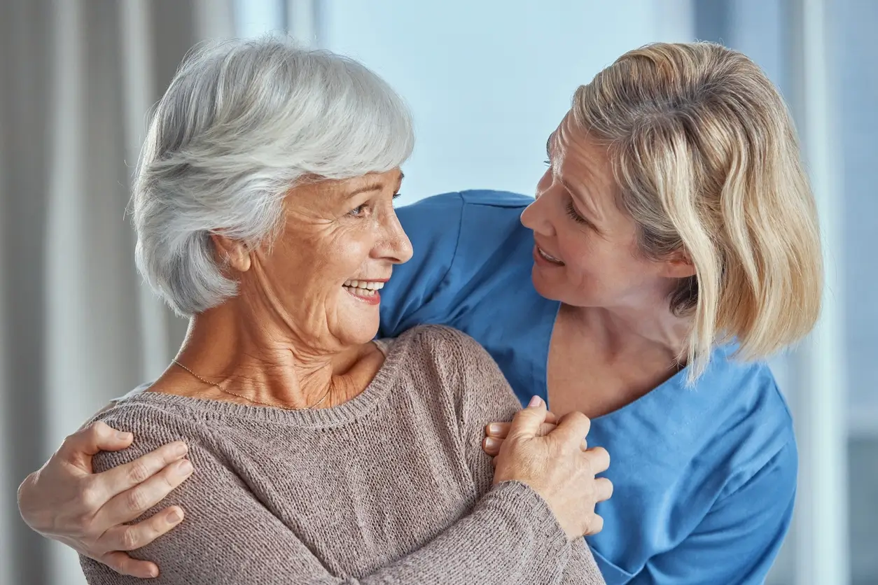 Mulher idosa sorri enquanto é abraçada por sua filha mais jovem na faixa dos 30 anos, ambas trocando um olhar carinhoso em um ambiente iluminado
