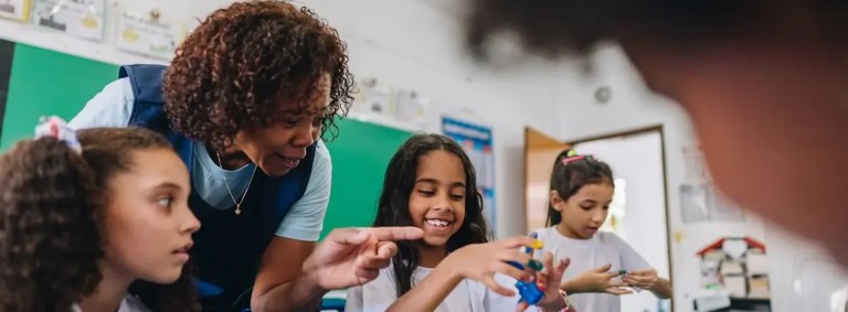 professora sorri enquanto orienta um grupo de meninas envolvidas em uma atividade lúdica com peças de montar