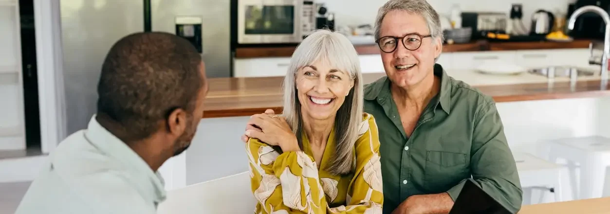 Casal de meia idade sentado à mesa sorri calorosamente para um homem mais jovem, em um ambiente de cozinha iluminado e moderno