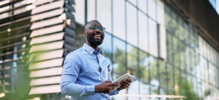 Homem de negócios sorridente segurando um tablet ao ar livre em frente a um prédio de vidro, representando sucesso na carreira ou negócios