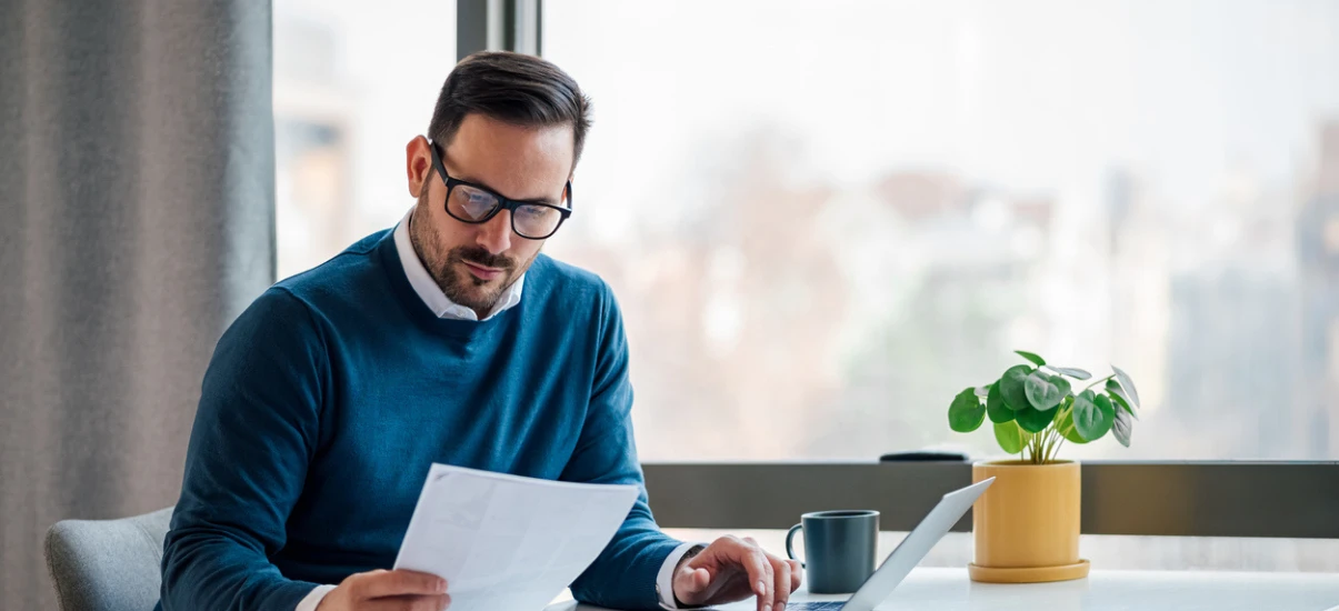 Homem de negócios concentrado analisando documentos em escritório moderno com iluminação natural, ao lado de uma planta verde e uma caneca.