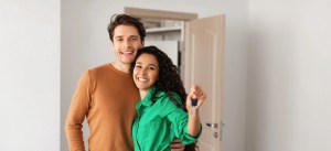 Casal feliz e sorridente posando para foto em casa, mulher com roupa verde e homem com camisa marrom, segurando uma chave na mão.