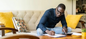 Homem estudando ou trabalhando em casa, sentado em um sofá com almofadas amarelas e utensílios de escritório, usando óculos e vestindo uma camisa azul escura.