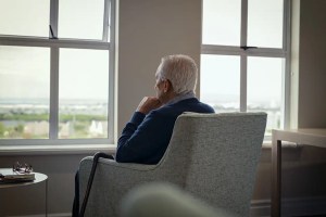 Homem idoso com cabelo branco sentado em uma cadeira observando a vista de uma janela grande, pensando ou refletindo, em ambiente de escritório ou residência com iluminação natural.