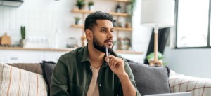 Homem pensativo sentado no sofá em casa, com expressão de reflexão, decoração moderna e iluminação natural na sala.