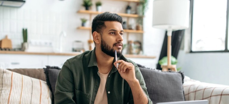 Homem pensativo sentado no sofá em casa, com expressão de reflexão, decoração moderna e iluminação natural na sala.